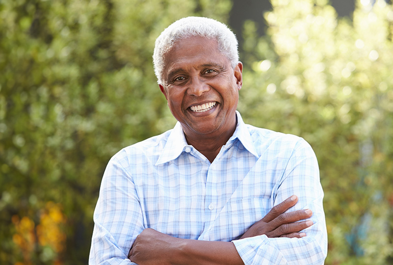The image shows a smiling man with gray hair, wearing a light blue button-up shirt, standing outdoors with a cheerful expression.