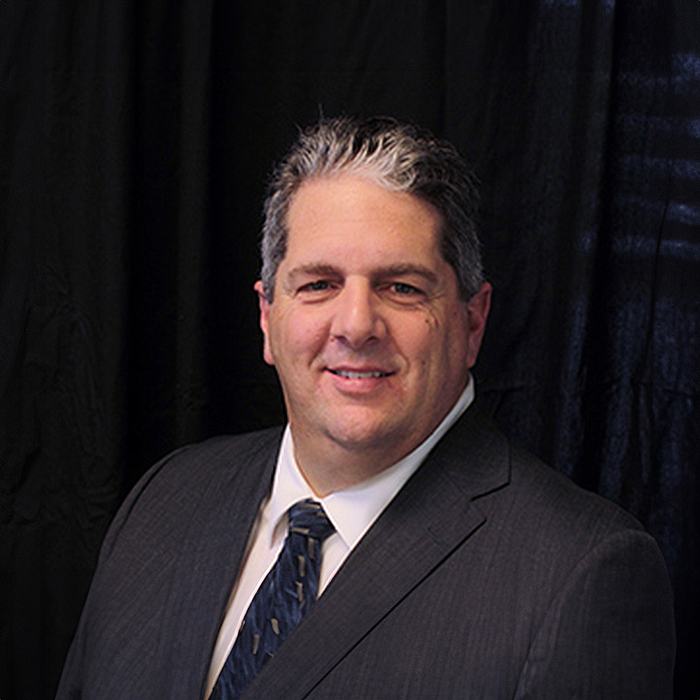 The image shows a man standing against a dark background. He appears to be wearing a suit with a tie, and he has short hair.
